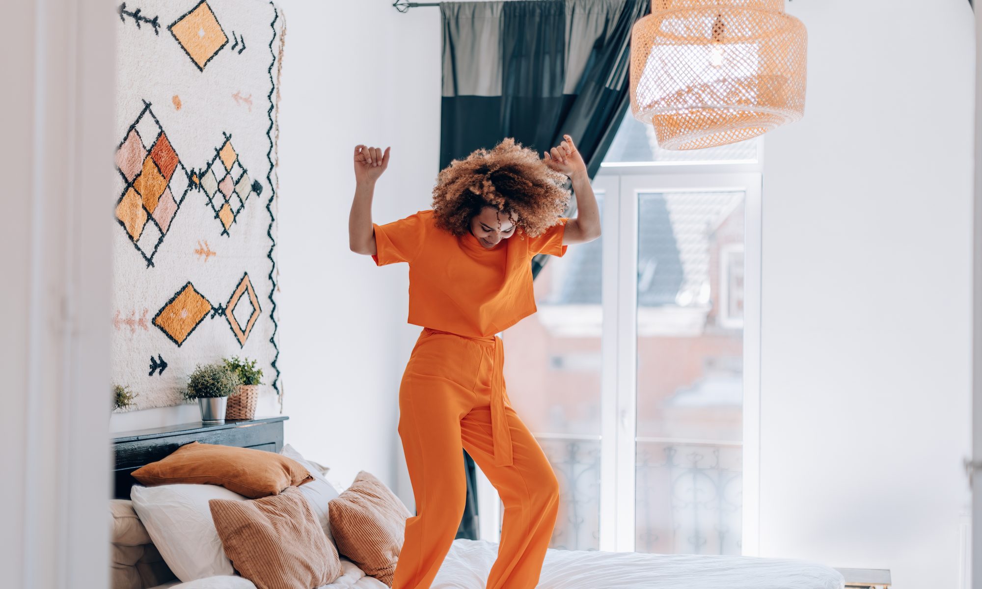 Happy Young Woman In Orange Outfit Dancing On Bed